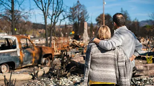 The back of a couple embracing in front of a disaster damaged home and vehicle.