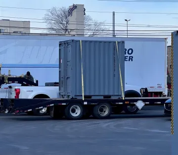 RAPID system. A large, corrugated metal container sitting on a flatbed trailer in a parking area in front of a pickup and a semi-truck.