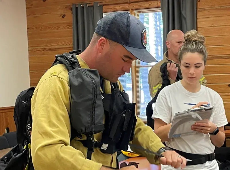 : In a wood paneled conference room a first responder wearing firefighting gear and a gray baseball cap, tightens a black Wildland Firefighter Respirator harness, while a data collector writes notes on a clipboard. In the background another firefighter lifting a backpack.