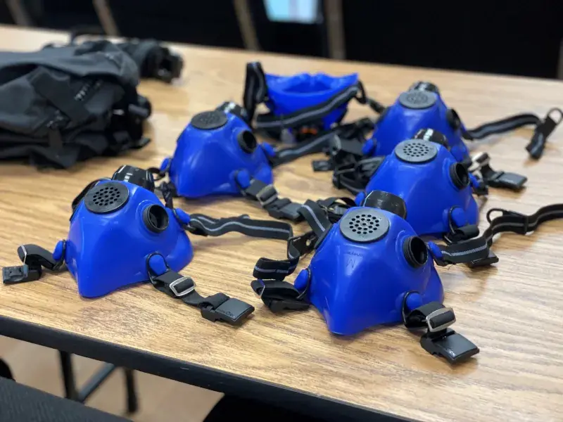 Six blue Wildland Firefighter Respirator half face masks lay lined up on a table. Background, a medium sized black nylon carrying pouch lays on the table behind the masks.