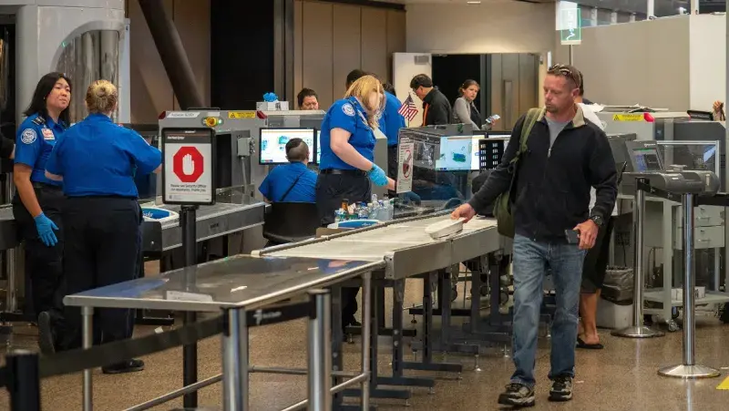 Travelers, including a man in a black jacket, blue jeans, and a bag over his shoulder, are passing through an airport security checkpoint. Personnel are manning their posts and viewing monitors, as carry-on items pass through scanners.