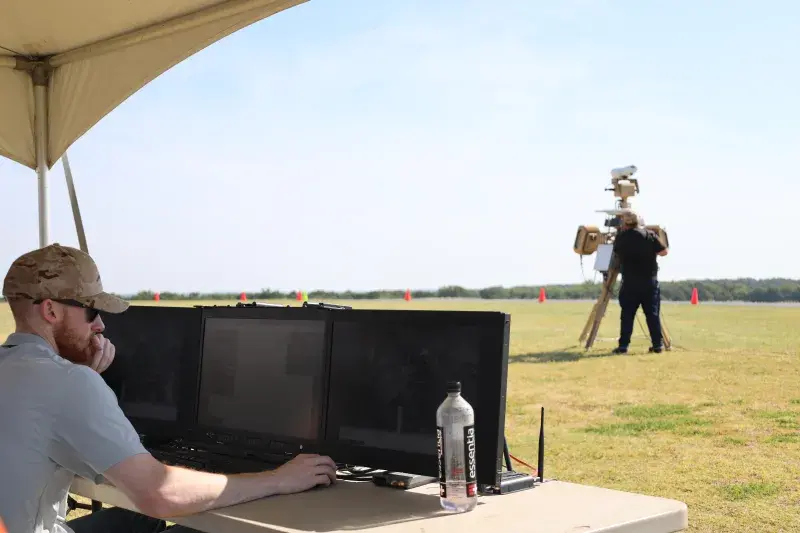 At right, a technician aims a drone detection system on a grass field while a second technician on the left sitting at a table scans monitors for any drone activity in the area during a demonstration in Oklahoma.