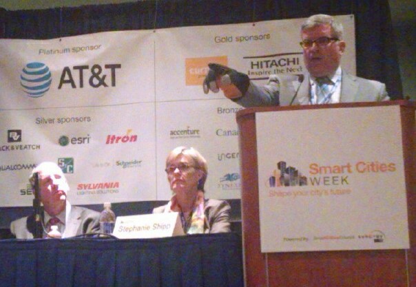 First Responders Group (FRG) Director Dan Cotter addresses a panel at Smart Cities Week convention in Washington, D.C. The background sponsor banner states: Platinum sponder, AT&T, Silver sponsers, CK & VEATCH, esri, Itron, llalcomm, src, SVLVANIA, Schgeneider, SE, Gold sponsors, Accenture, Canada, Gen, Bronz, HITACHI, Inspire the Next, Cur. Smart Cities Week, Shape your city’s future