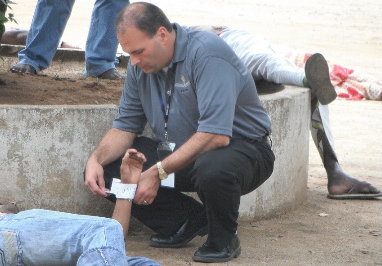 Fire Chief Robert McLafferty outside, kneeling and looking at a tag attached to a wounded victim laying on the ground.