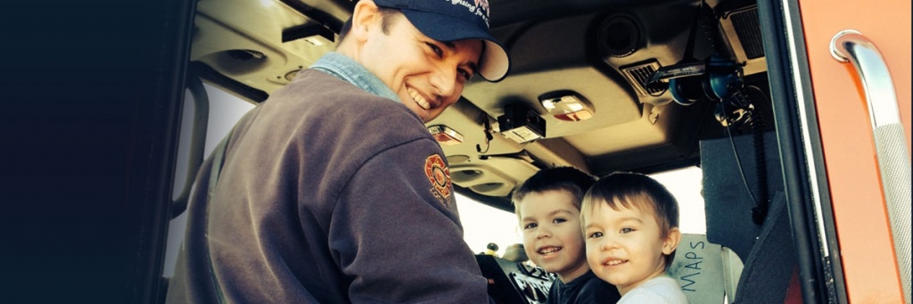 First Responder Jason Smith stands in front of two young boys smiling sitting in the front seat of an ambulance.