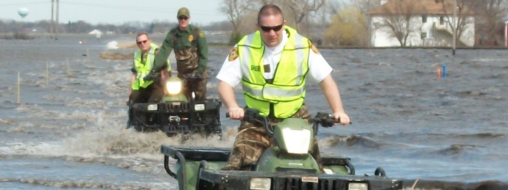 Photo of Paul D. Laney (front) on a four wheeler and two other responders behind him also on four wheelers, all riding through the water.