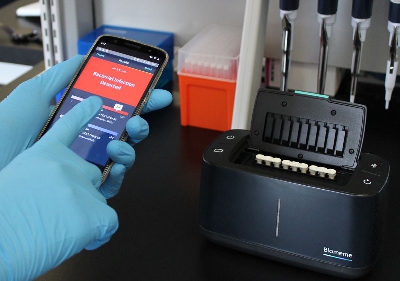 A researcher points to a result reading “Bacterial Infection Detected.” The black Franklin™ thermocycler by Biomeme, Inc. (right) can measure any DNA or RNA that Predigen, Inc. designs a test for. Inside the thermocycler are white test tubes with reagents to detect host response signature in samples. It can be operated by a smartphone app and can export data to the cloud.  Photo by Biomeme/Predigen.