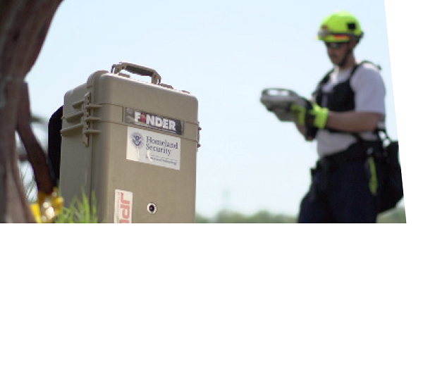 Close up of "FINDER" device, a beige case with "FINDER" and "DHS S&T logo" stickers on the front. Man in background with yellow hardhat is looking at device in his hands.