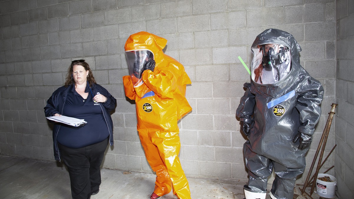 As an evaluator looks on and captures feedback, a pair of first responders, wearing personal protective equipment and in-suit communications gear, conducts a building entry drill.