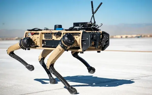 A tan, black and silvery 4-legged metallic robot dog is walking across grey cement pavement. Low tan-colored buildings are blurry but visible in the distance. In the extreme distance is a mountain range. The shadow cast by the robot dog indicates it is waking away from the Sun.