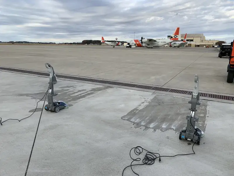 On a concrete runway, there are 2 vacuum machines and wet patches in front of them. Cords a trailing behind the vacuums. A brown grate is behind the vacuums, and in the background can be seen several Coast Guard airplanes and buildings. The sky is overcast. 