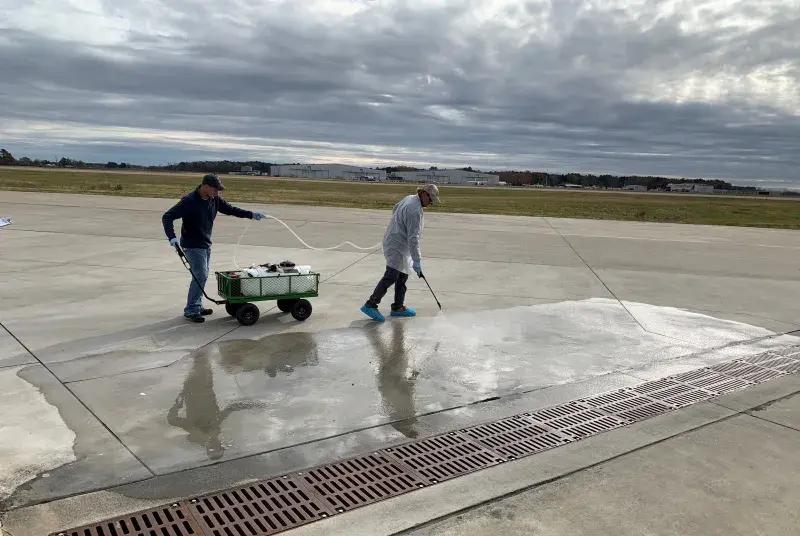 Two male EPA researchers spray water on concrete runway with a brown grate in front and green grass, airplanes, airplane hangars in the background and a sky overcast with grey clouds. researchers from EPA spray water on concrete. The men’s reflection can be seen in the sprayed concrete. The first man is dressed in white lab coat, white baseball cap, the second is dressed in navy coat and baseball cap and blue jeans.