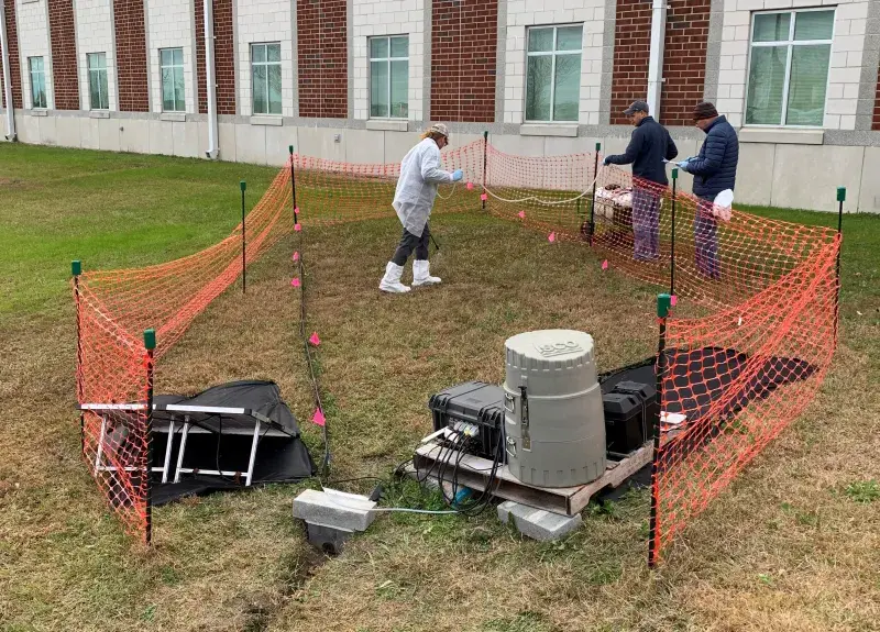 Three male EPA researchers  work on a grassy site next to an office building. The site is surrounded by a yard tall orange netting. In side is a variety of sensor and sampling technology. Inside the fence is a man dresses in white lab coat and white booties, who is sprays the grass. Two men outside the far end of the nefence are dressed in navy – on is holding the hose of a rolling sprayed and the other is recording something on a pad. At the front, there is an opening in the fence, where there is a narrow ditch.