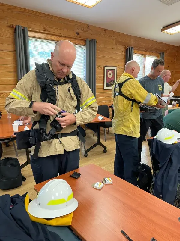 In a wood-paneled conference room a first responder wearing a beige firefighters protective jacket and a black Wildland Firefighter Respirator harness adjusts the water bottle-sized WFFR filter unit. A white firefighter helmet is on a table in front of them. In the background are other firefighters and data collectors preparing gear.