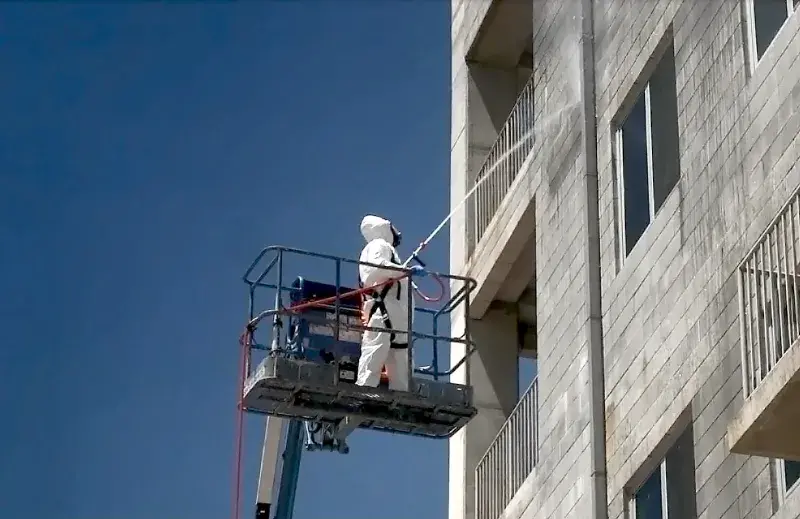 A man dressed in white Tyvek protective overalls with a hood wears a breathing mask. He is spraying the wall of the 4-5th floor of a tall grayish building while standing on raised vehicle platform in the background of a blue sky. 