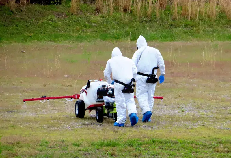 Two men with their backs toward us, one shorter than the other, push a wheeled container full of liquid. A red horizontal tube attached to the container sprayers the liquid from multiple places on a grass tube lawn. The men are dressed in white Tyvek overalls, wear breathing masks and black belts holding apparatuses that push air to the masks and wear blue gloves and blue plastic over their shoes.