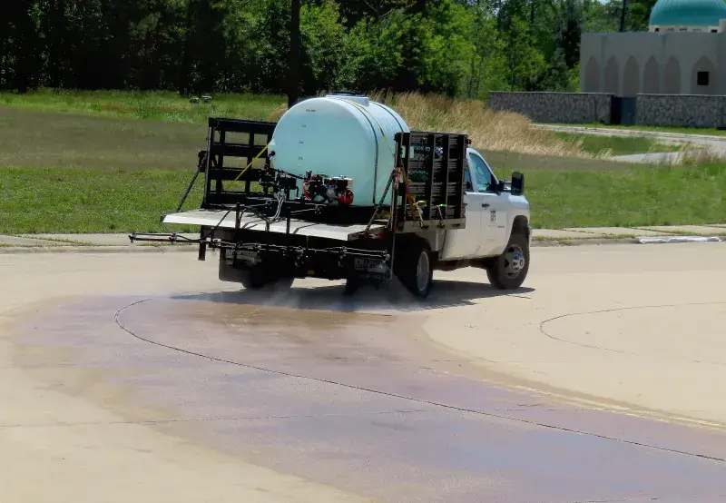 A truck sprays decontamination liquid using a boom sprayer - a horizontal black pipe at the back end of the truck, from which pipe multiple nozzles spray the liquid on the concrete road, leaves a wide wet trail in its wake.  The truck caries a cylindrical horizontal container which is secured by ropes. In the background could be seen a lawn, a concrete building with turquoise dome. 