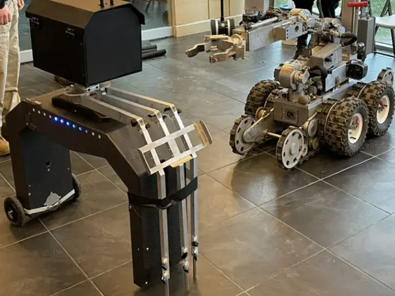 Xoran’s 3D X-ray scanner stands in a room, on a tiled floor. Illuminated blue lights on top of the scanner indicate that it is ready for use. To the right of the scanner is a six-wheeled remote-controlled robotic truck with a crane-shaped arm, preparing to pick up the X-ray.