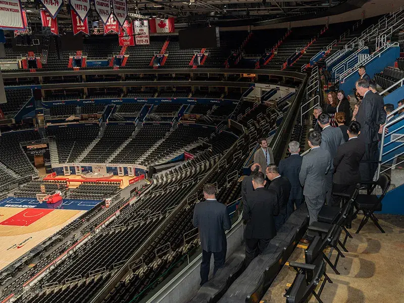 A group of officials are seen from behind standing up in the stands of the Capital One Arena in Washington, DC during a tour, looking down at a basketball court below. 