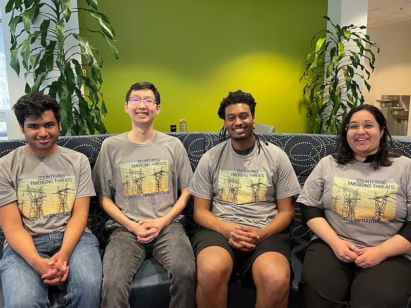 Four Criminal Investigations and Network Analysis Center/George Mason University students in matching t-shirts that say “Countering Emerging Threats toCritical Infrastructure,” sit on a blue-patterned couch as they pose for a photo.