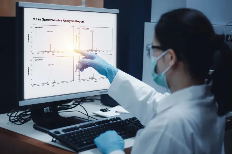 A female scientist, wearing a white lab coat and a mask, is pointing at a “Mass Spectrometry Analysis Report” on a computer screen. Four samples are in individual graphs on the computer screen. The chemical signatures of the compounds are revealed by the spikes in the graphs.