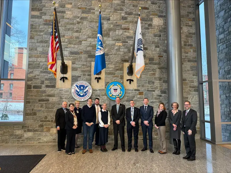 Under Secretary Kusnezov poses with members from the S&T and UK delegations in front of the US, DHS, and US Coast Guard flags at the St. Elizabeths Campus.