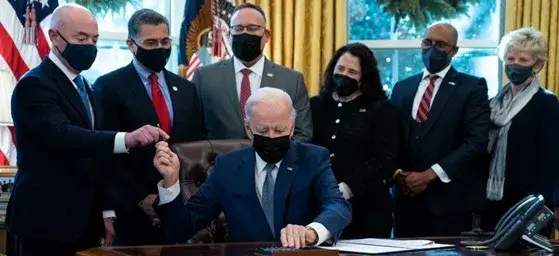 President Biden hands Secretary Alejandro Mayorkas a pen during the signing of Executive Order 14058, "Transforming Federal Customer Experience and Service Delivery to Rebuild Trust in Government." A group of other people stand behind President Biden, and next to Secretary Mayorkas. All are wearing masks.