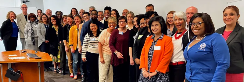 A large group of workshop participants lined up and smiling for the camera in a meeting room with a desk holding a microphone and a whiteboard in the background. 