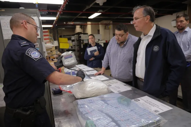 From right: Dr. Kusnezov, Adam Cox, and Steve Feder look on as CBP Officer demonstrates a handheld optical sensor used to identify bulk drugs in international mail shipments.