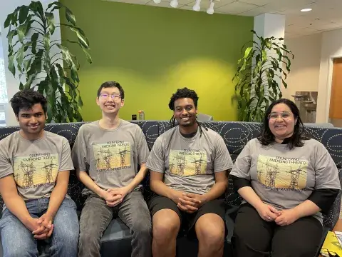Four Criminal Investigations and Network Analysis Center/George Mason University students in matching t-shirts that say “Countering Emerging Threats to Critical Infrastructure,” sit on a blue-patterned couch as they pose for a photo. 