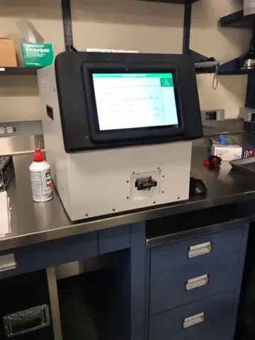 The mass spectrometer is a metal box about 18 inches square. It is on top of a desk or counter. Half of the front of the spectrometer is a computer screen facing us. There are other objects on the counter, like a computer mouse, rubber gloves, cleaning wipes, and an air spray can for cleaning surfaces.