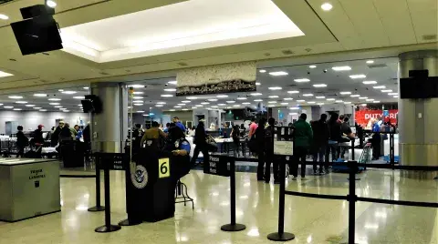 Transportation Security Administration (TSA) checkpoint at an airport. A TSA employee is seated at the gate. Multiple lines of passengers are putting their belongings on conveyor belts to be scanned.