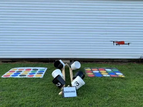 Drone flying over a sheet with different colored circles on it, a cluster of buckets labeled “ground”, and another sheet with colored circles.