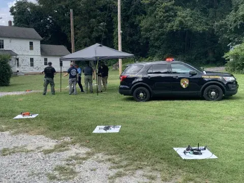 Three white squares lined up on the grass, each with a target and a drone landed upon it. Black SUV and tent in background with test participants beneath it.