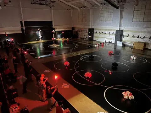 People standing on the side and drones being flown inside of a darkened gymnasium with spotlights here and there, and clusters on buckets arranged neatly on the ground and on wooden posts.