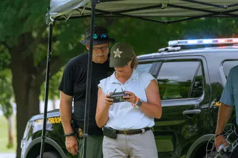 A female drone operator being observed by a male operator during testing standing under a tent near a state trooper vehicle. by test proctor