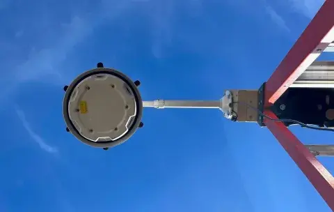 View from below of the circular white gunshot detection technology. The technology is mounted via a rod to the right on a red ladder and only the top of the ladder is showing.