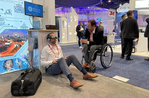 A man sitting on the floor wearing virtual reality goggles and a man in a wheelchair at the S&T booth at CES