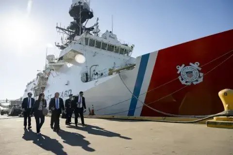 Homeland Security Secretary Alejandro Mayorkas tours the U.S. Coast Guard Cutter Stratton at the Coast Guard Island Alameda, California. He is walking with a group of people down a dock with a ship in the background.