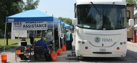 A white bus FEMA parked next to blue pop-up shelters, with SBA Disaster Assistance banners. There are people seated and standing next to tables with computers.