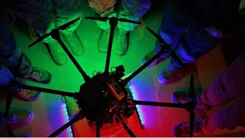 Six pairs of feet in combat boots and hiking shoes surround a small unmanned aircraft system resting on the ground that is emitting green, purple, blue and red light.