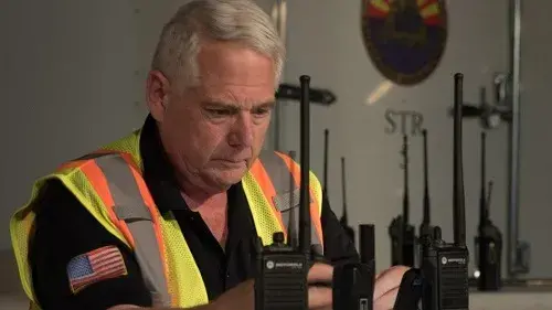 Man reading screen of cell phone on table while holding a handheld radio in right hand.