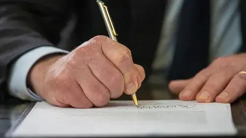 A man's left hand holds a piece of paper with his left hand and signs it with his right with a gold pen. The man's face is not visible nor is the writing on the page.