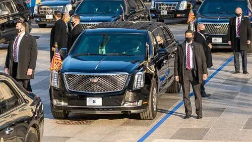 The Presidential limousine is flanked by 6 Secret Service officers in suits, black jackets, and COVID masks.