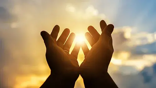 Hands, cupped together as if in worship, with sunlight and a cloudy sky behind them.