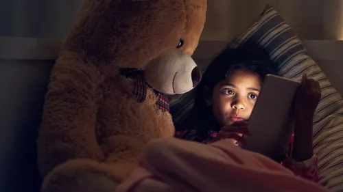 A photo of a little girl and a large teddy bear looking at a tablet in the dark.