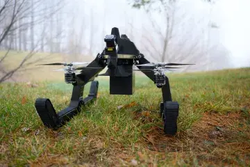 Pegasus III Transformable Air-Ground Robotic System drone on the ground moving along a field of short grass on it’s treads. In the background – a grassy hill and a few trees. 