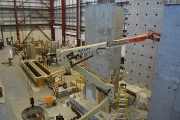 An expansive space view from upper region depicts the enormous structural engineering laboratory at the University of Kansas. The space is as large as a several-story building and resembles an airplane hangar. There are various building instruments, tools and materials as commonly seen on construction sites.