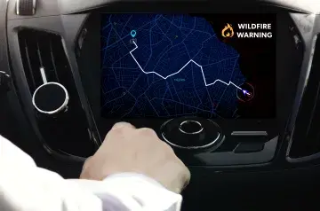 Representation of an infotainment console in a car that shows a navigational mapping app. The digital map on the infotainment console is showing directions to route a driver around a fire incident. The danger zone is marked with a fire symbol and the words “Wildfire Warning.” The driver’s hand is visible and might be reaching for the console.