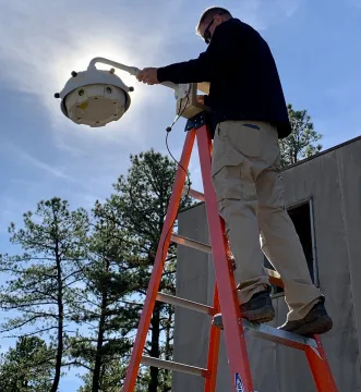 A man stands on a ladder installs the circular/round SDS gunshot Detection technology .